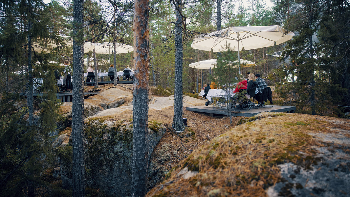 Ravintolapöydät syksyisen metsän keskellä, pöydissä istuu ihmisiä